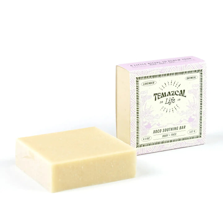 Temazcal Life Xoco Soothing Bar shown unboxed next to its lavender-themed packaging, highlighting its simple, creamy color and square shape.