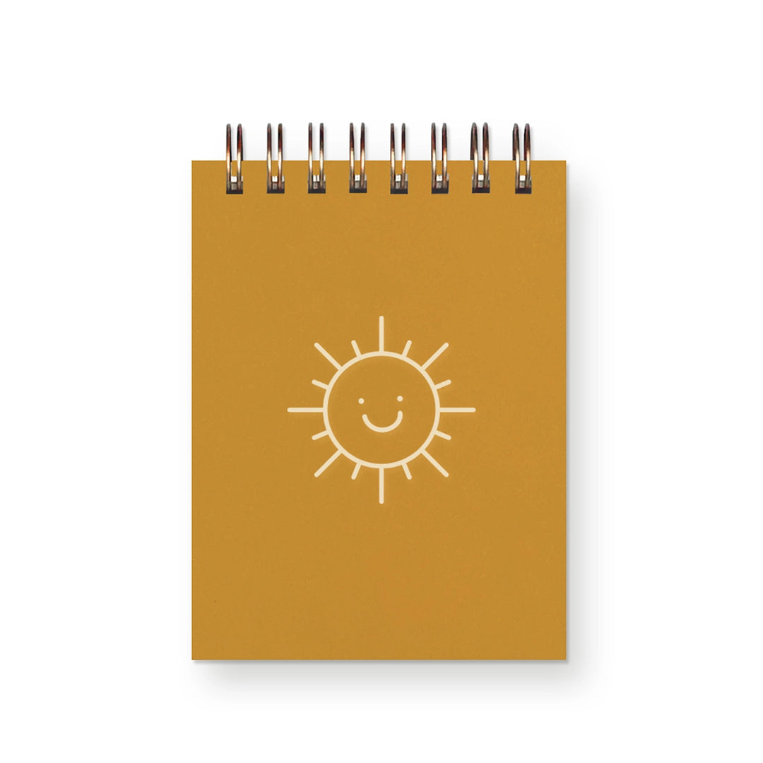 Small spiral notebook with a mustard yellow cover and a simple, smiling sun illustration in white.