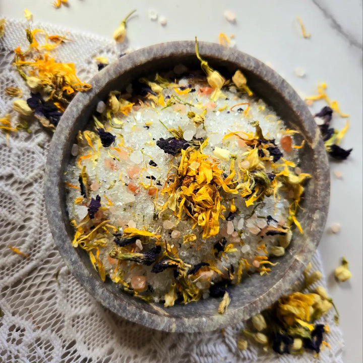 A close-up of dried flowers and bath salts in a small stone bowl, surrounded by scattered petals.