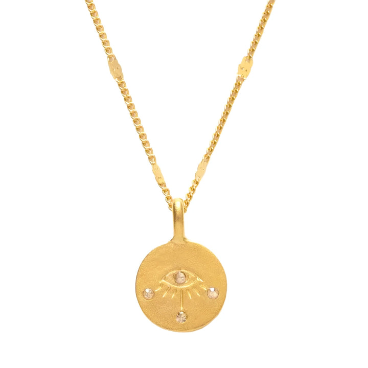 Close up View of a gold Eye of Protection Necklace featuring a round pendant with an engraved eye design and crystal accents. The pendant hangs from a delicate gold chain with subtle decorative links,