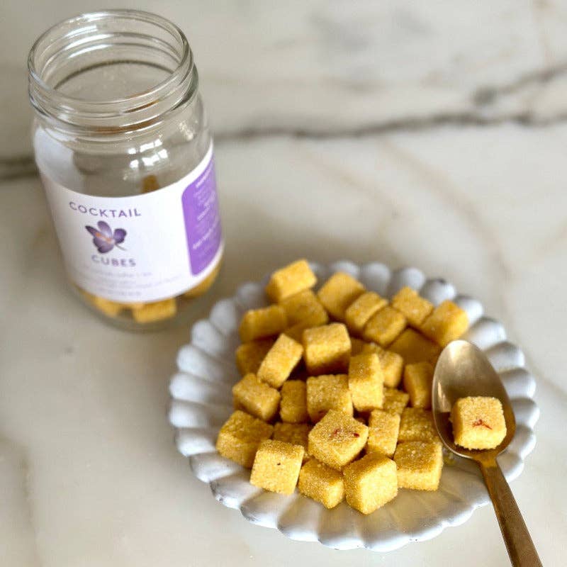 white plate with sugar cubes and a gold spoon with an open jar of the sugar cubes in the background.