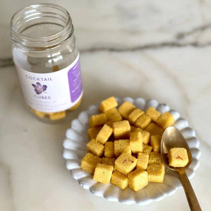 white plate with sugar cubes and a gold spoon with an open jar of the sugar cubes in the background.