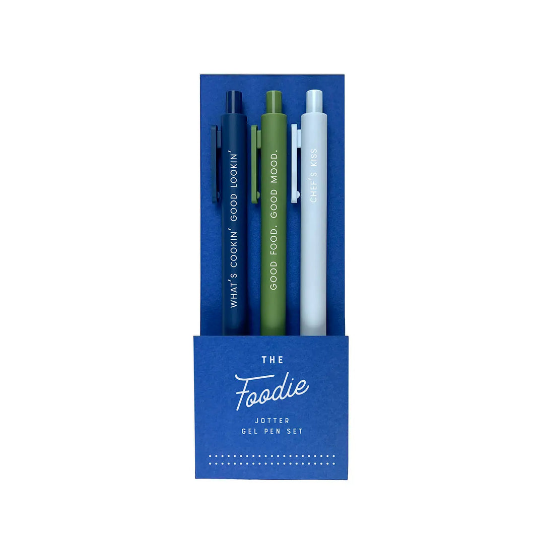 A set of three colorful gel pens in a blue box labeled "The Foodie." Each pen has a fun food-themed phrase printed on it, including “What’s Cookin’ Good Lookin’,” “Good Food. Good Mood.,” and “Chef’s Kiss.”