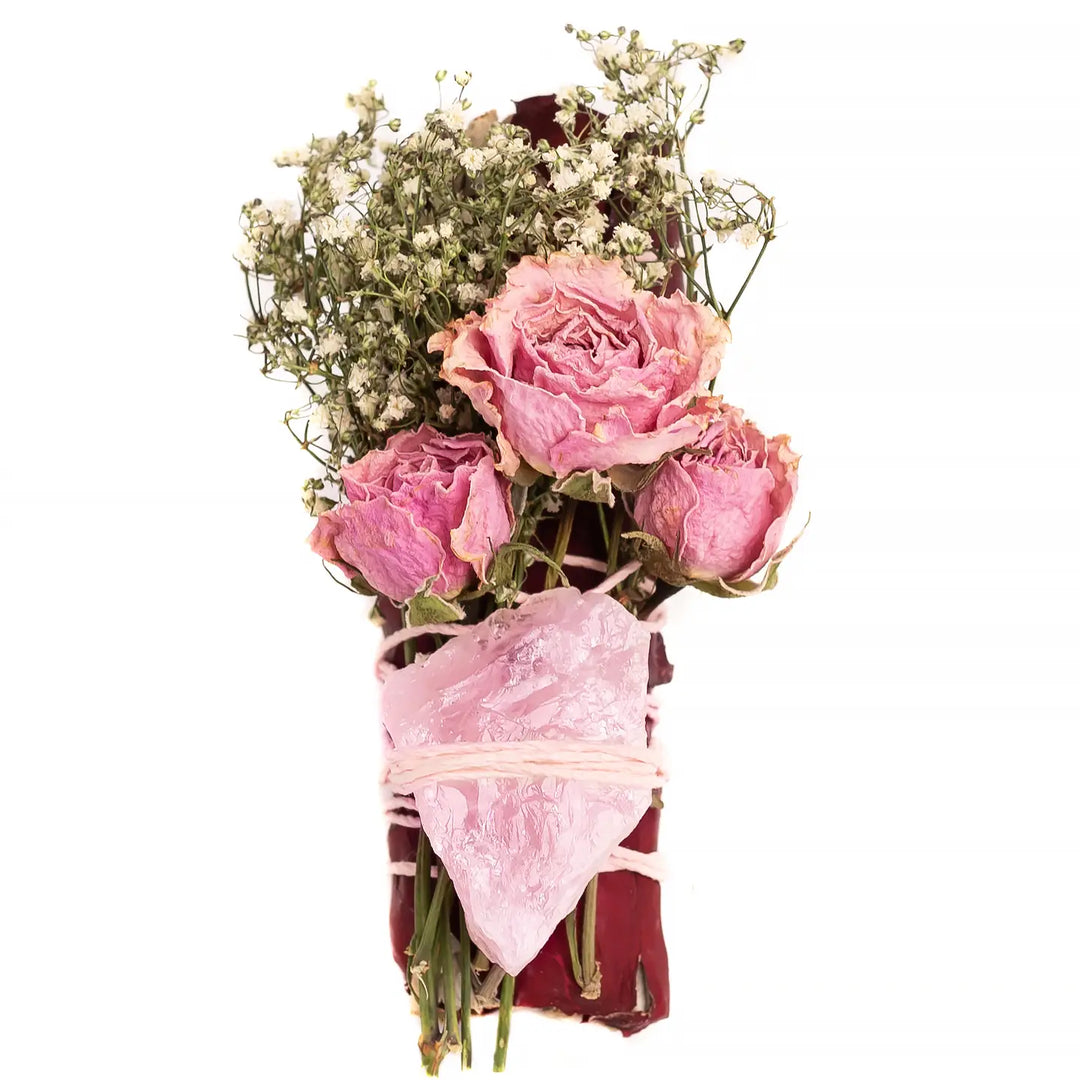 A smudge bundle wrapped in white twine, featuring dried pink roses, baby’s breath flowers, and a large raw rose quartz crystal tied on top sage leaves,
