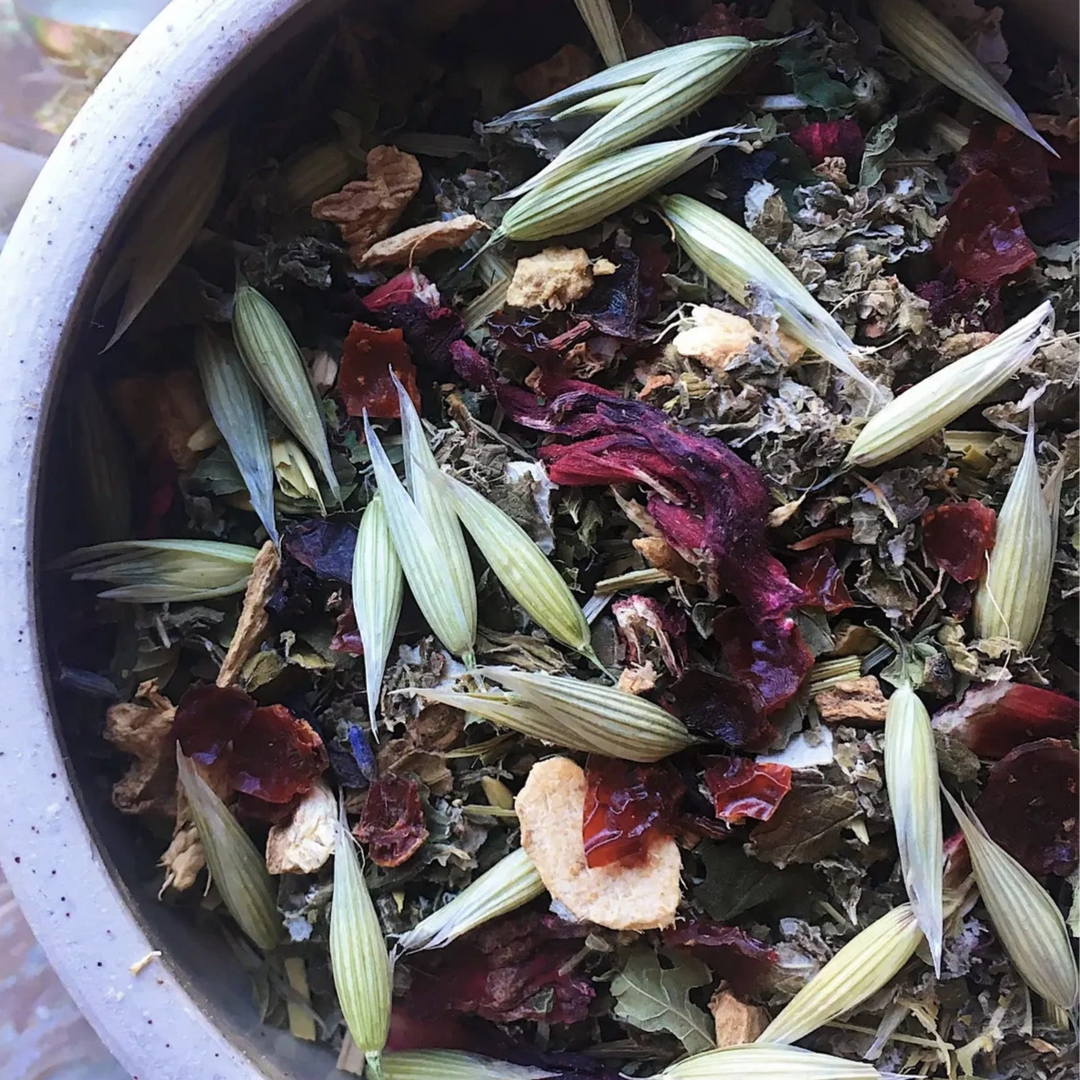 Ceramic bowl filled with Full Moon loose leaf herbal tea blend, featuring vibrant green oatstraw, rose petals, dried citrus, and herbs.