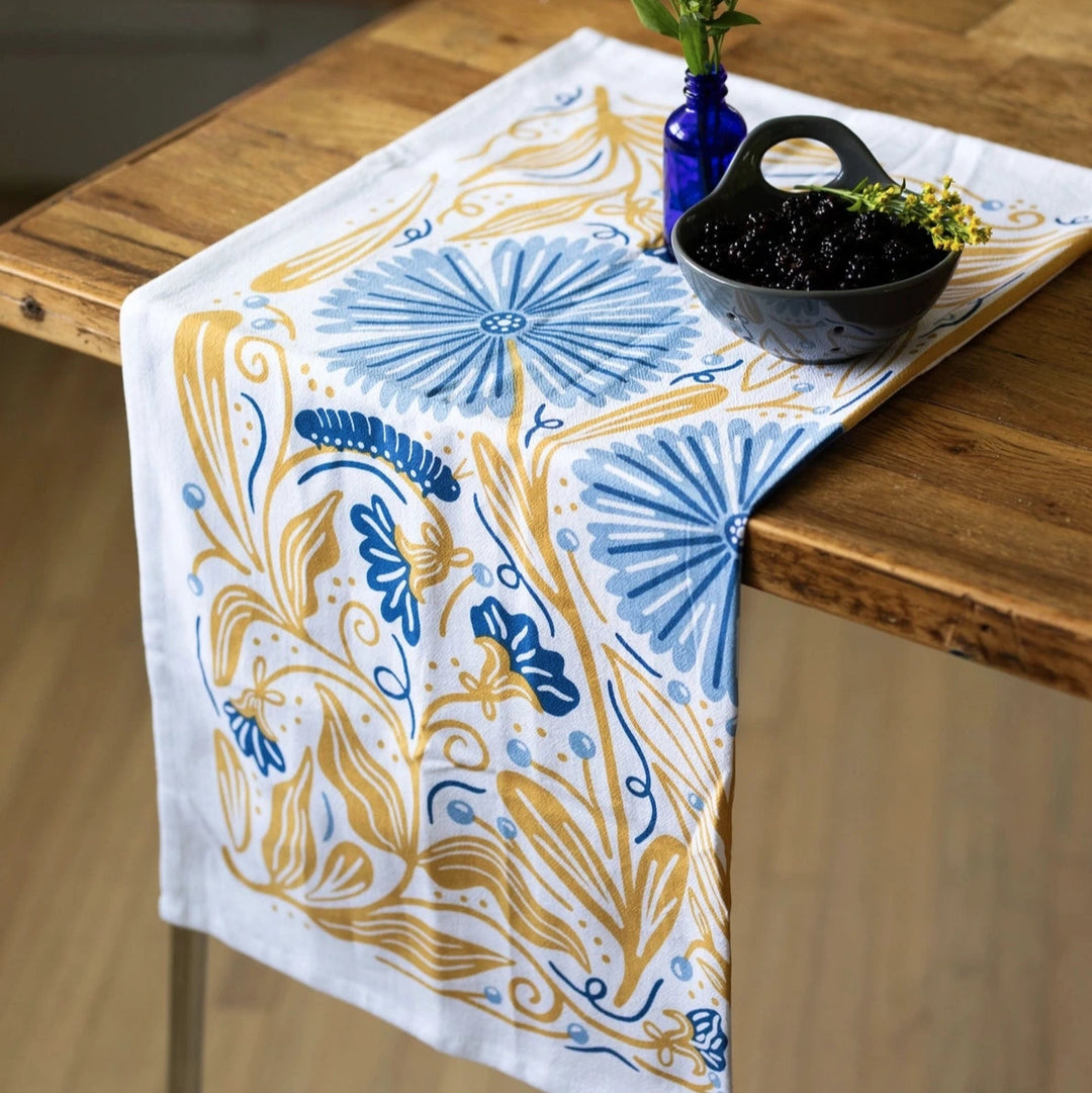 Colorful tea towel with a blue and gold floral and caterpillar design draped over the edge of a wooden table. A small blue glass vase with yellow flowers and a bowl of dark berries sit on top of the towel.
