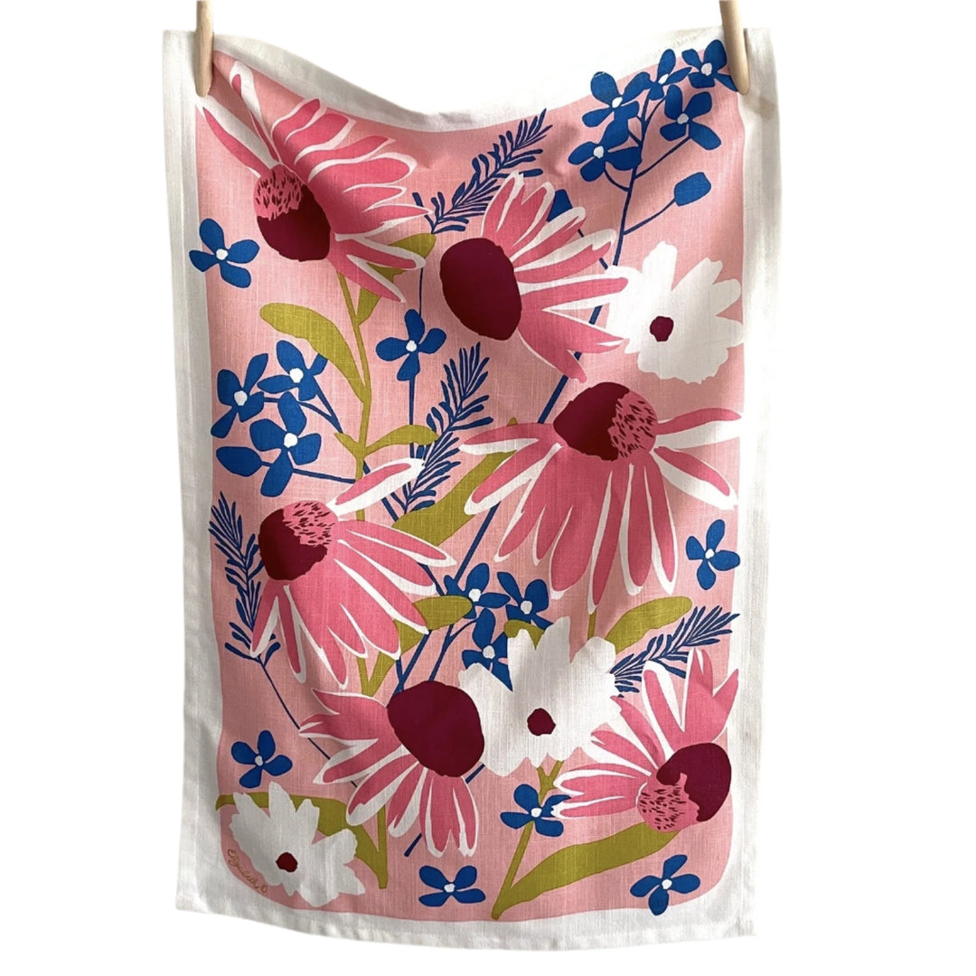 Echinacea-themed tea towel hanging on a line, featuring pink, burgundy, blue, and white flowers on a soft pink background.