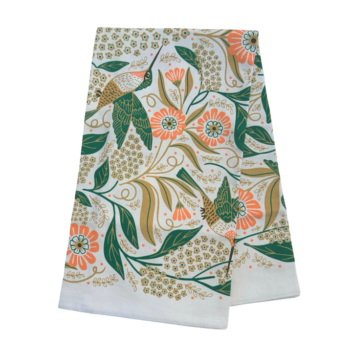 A White kitchen towel with a symmetrical floral and hummingbird design in shades of green, pink, and gold folded in quarters.