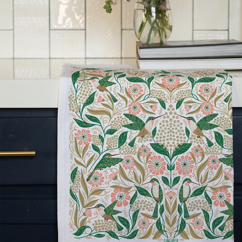Cotton tea towel draped over a dark cabinet, displaying a symmetrical floral and hummingbird design in shades of green, pink, and gold on a white background. A vase of fresh flowers sits nearby.