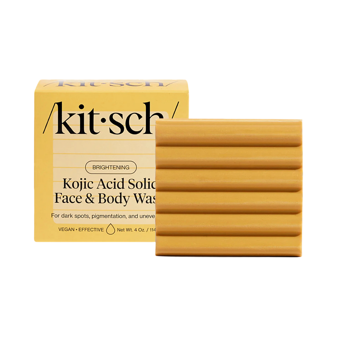 A Kitsch Kojic Acid Solid Face & Body Wash bar in a golden-yellow shade with a ridged texture, displayed next to its matching yellow packaging. The box highlights key benefits such as brightening, targeting dark spots, pigmentation, and uneven skin tone, and mentions that the product is vegan and effective.