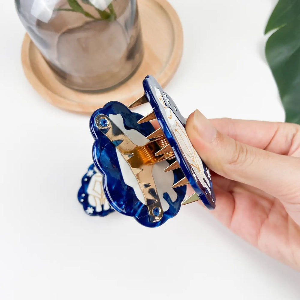 hand holding open a blue hair clip that features an outline of a woman in gold sitting down holding her knees in