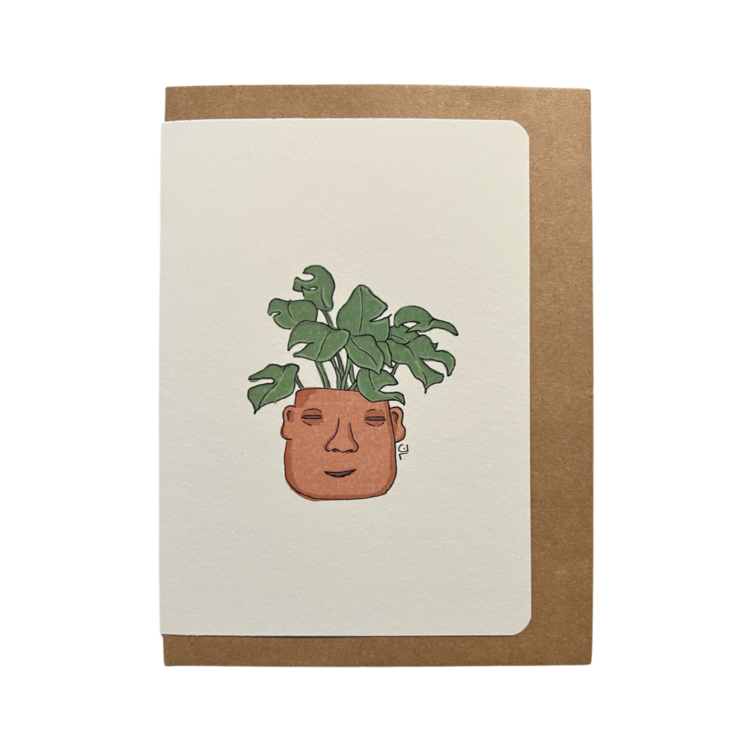 Illustration of a terracotta planter shaped like a serene face, adorned with lush green leaves, featured on a greeting card with a kraft envelope in the background.