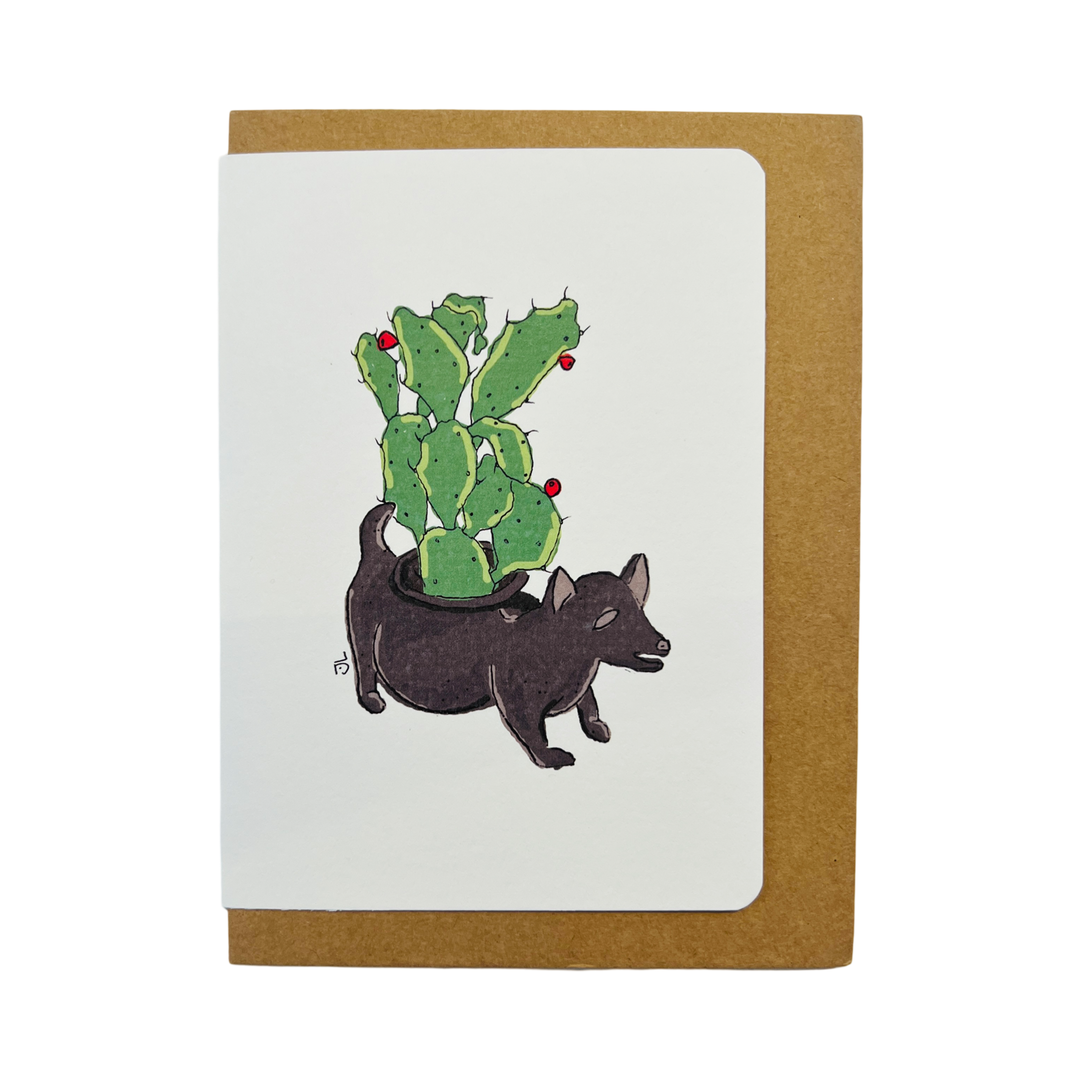 A greeting card featuring an illustration of a black dog-shaped planter filled with a green cactus adorned with red tunas, set against a clean white background with a brown kraft envelope.