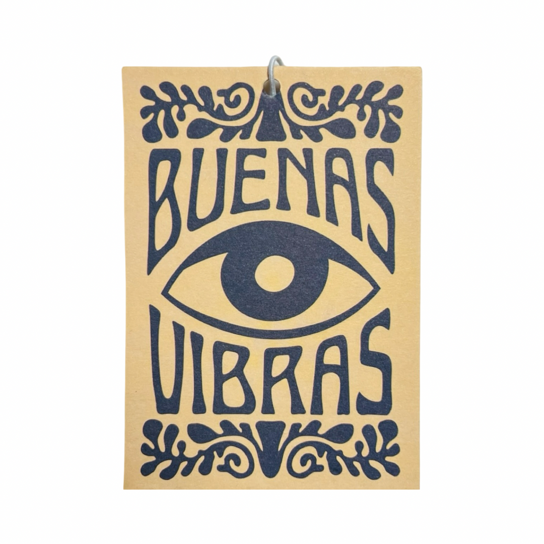 Rectangular air freshener with a tan background featuring bold black typography that reads "Buenas Vibras," a central mystical eye illustration, and decorative elements.