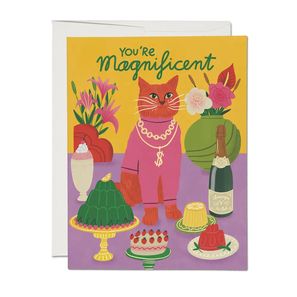 Greeting card with an illustration of a red-orange cat in a pink outfit and dollar-sign chain, surrounded by desserts, flowers, and champagne. Text reads “You’re Magnificent” at the top.