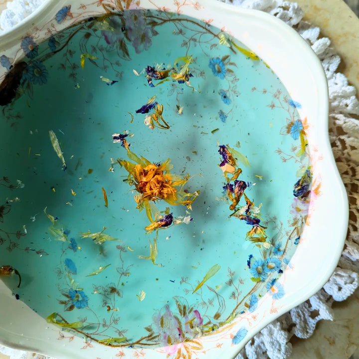 A floral botanical bath soak floating in light blue water inside a vintage floral dish.