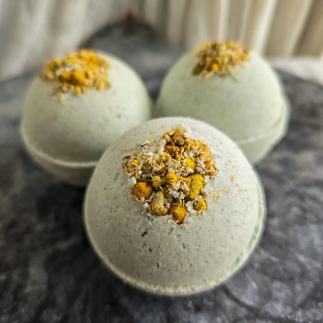 Three pale blue bath bombs topped with dried chamomile flowers on a dark stone surface.