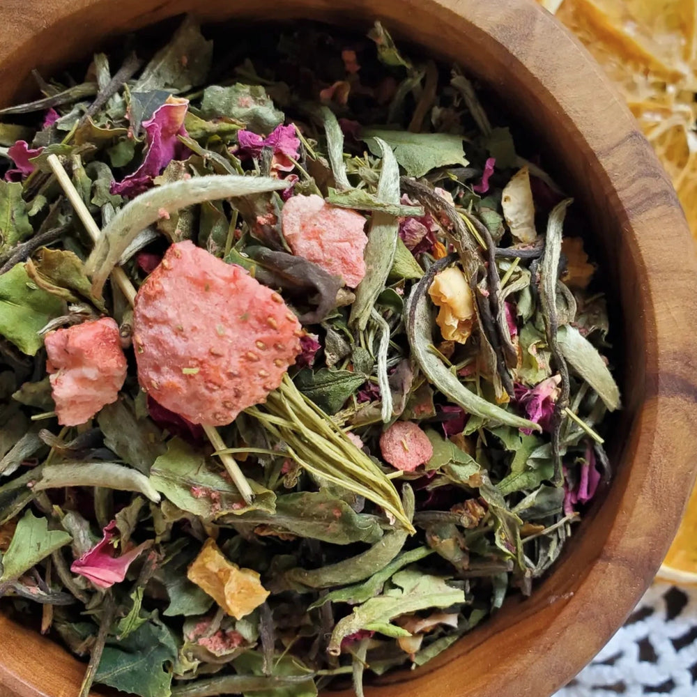 Close-up of a wooden bowl filled with loose Strawberry Sun tea blend, showing dried white tea leaves, strawberry pieces, and flower petals. Dried orange slices are in the background.