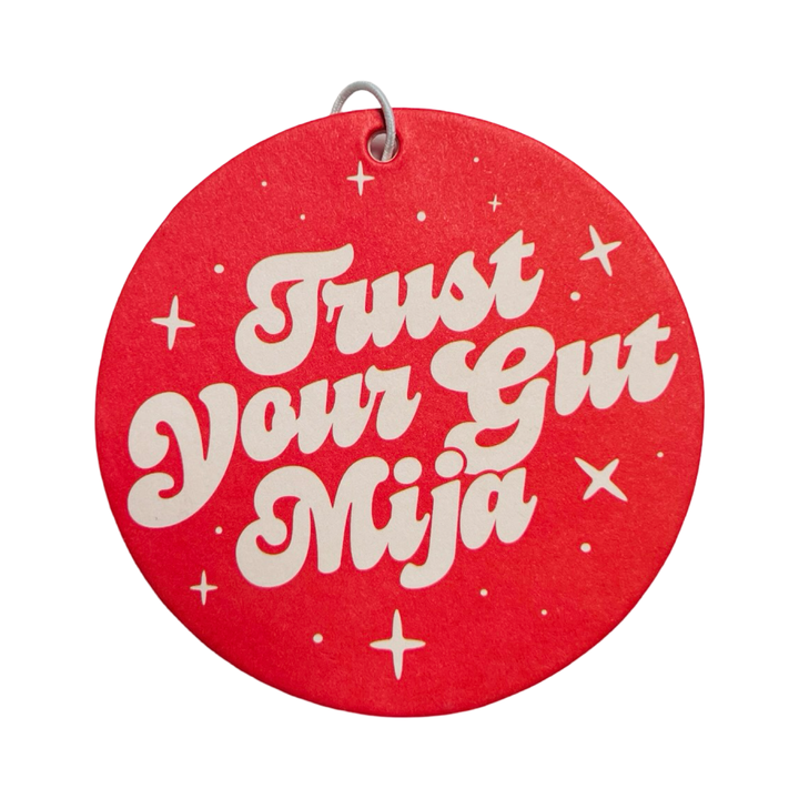 Round red air freshener with white retro-style text that reads "Trust Your Gut Mija," accented with small stars, infused with an orange scent.