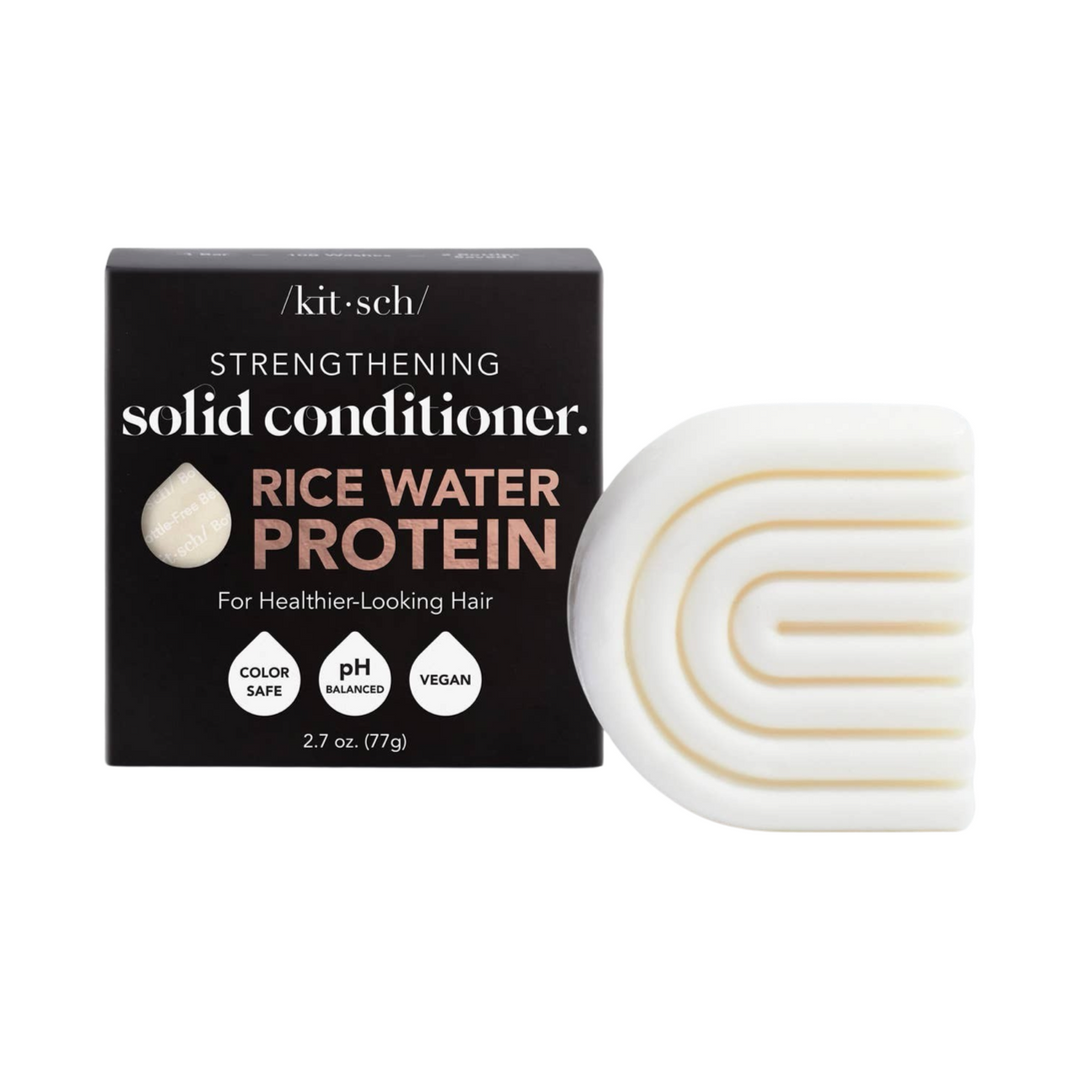 Black branded box of conditioner bar and at its side a cream colored bar of conditioner in the shape of a rainbow. Brand: Kitsch