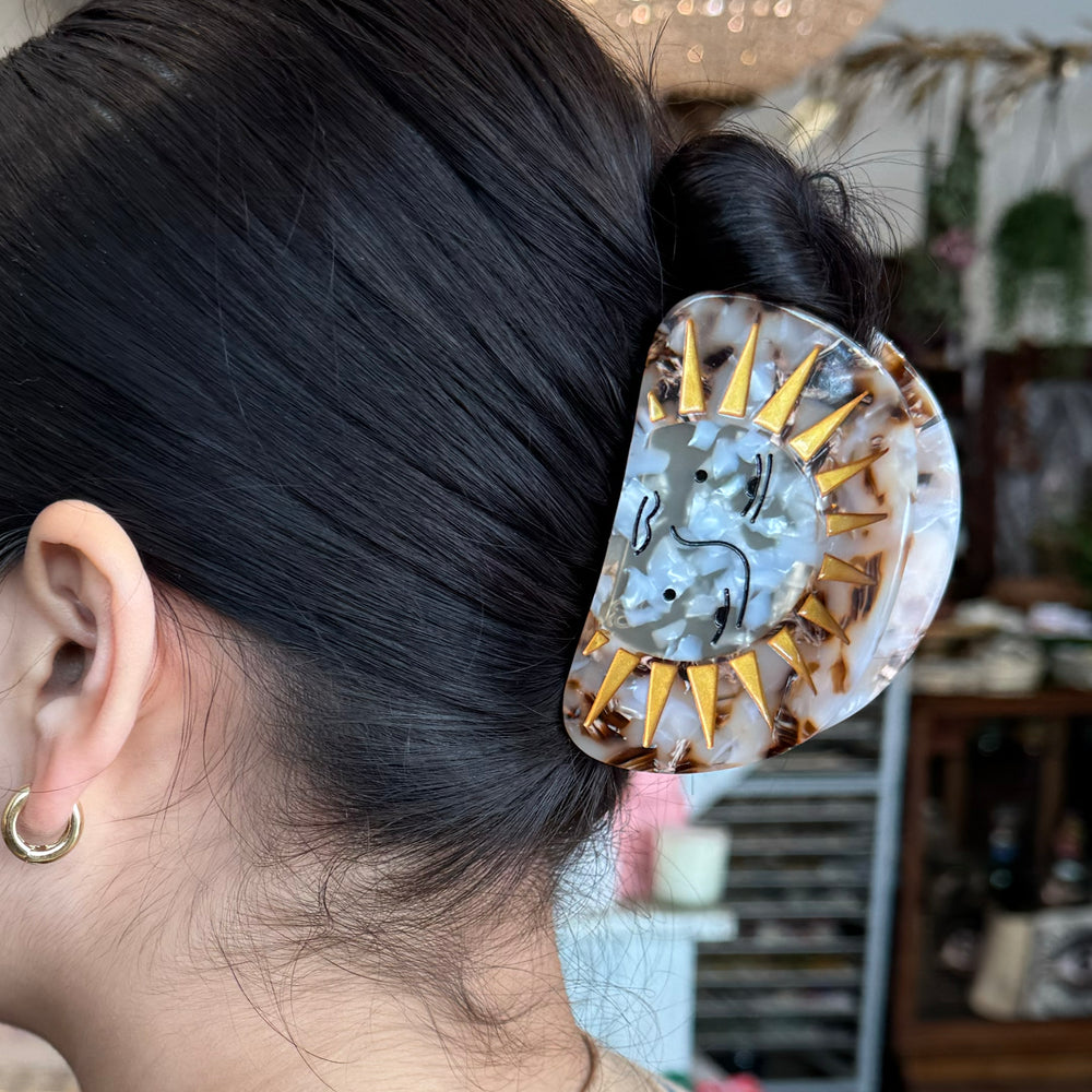 A sun-themed hair clip with a marbled finish, golden sun rays, and an artistic face design, set against a white background.