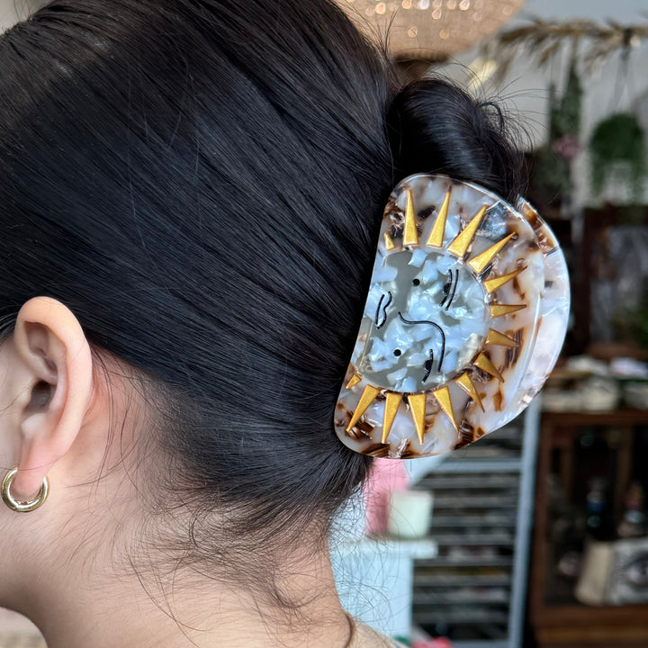 A sun-themed hair clip with a marbled finish, golden sun rays, and an artistic face design, set against a white background.