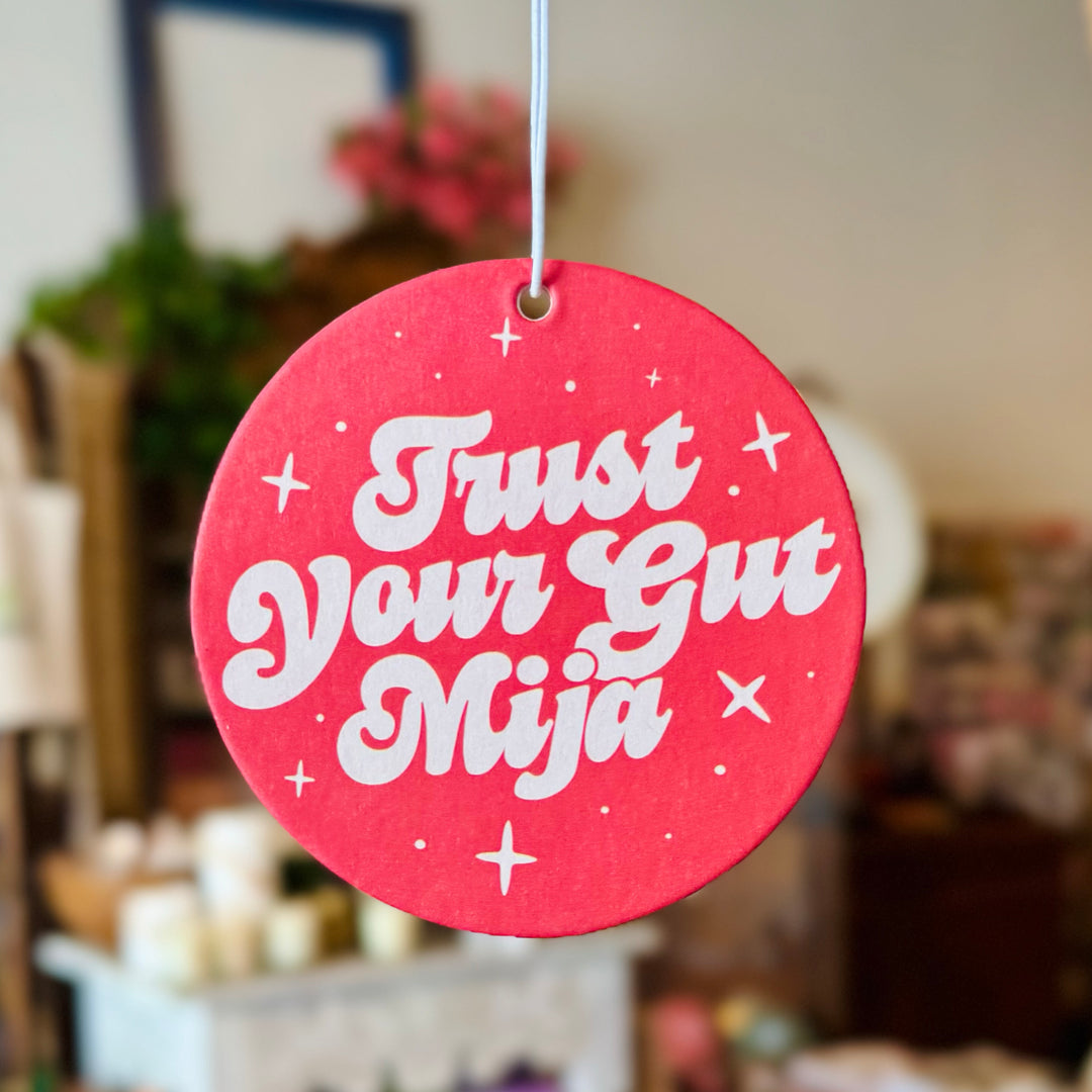 photo of round red air freshener with white retro-style text that reads "Trust Your Gut Mija," accented with small stars, infused with an orange scent.