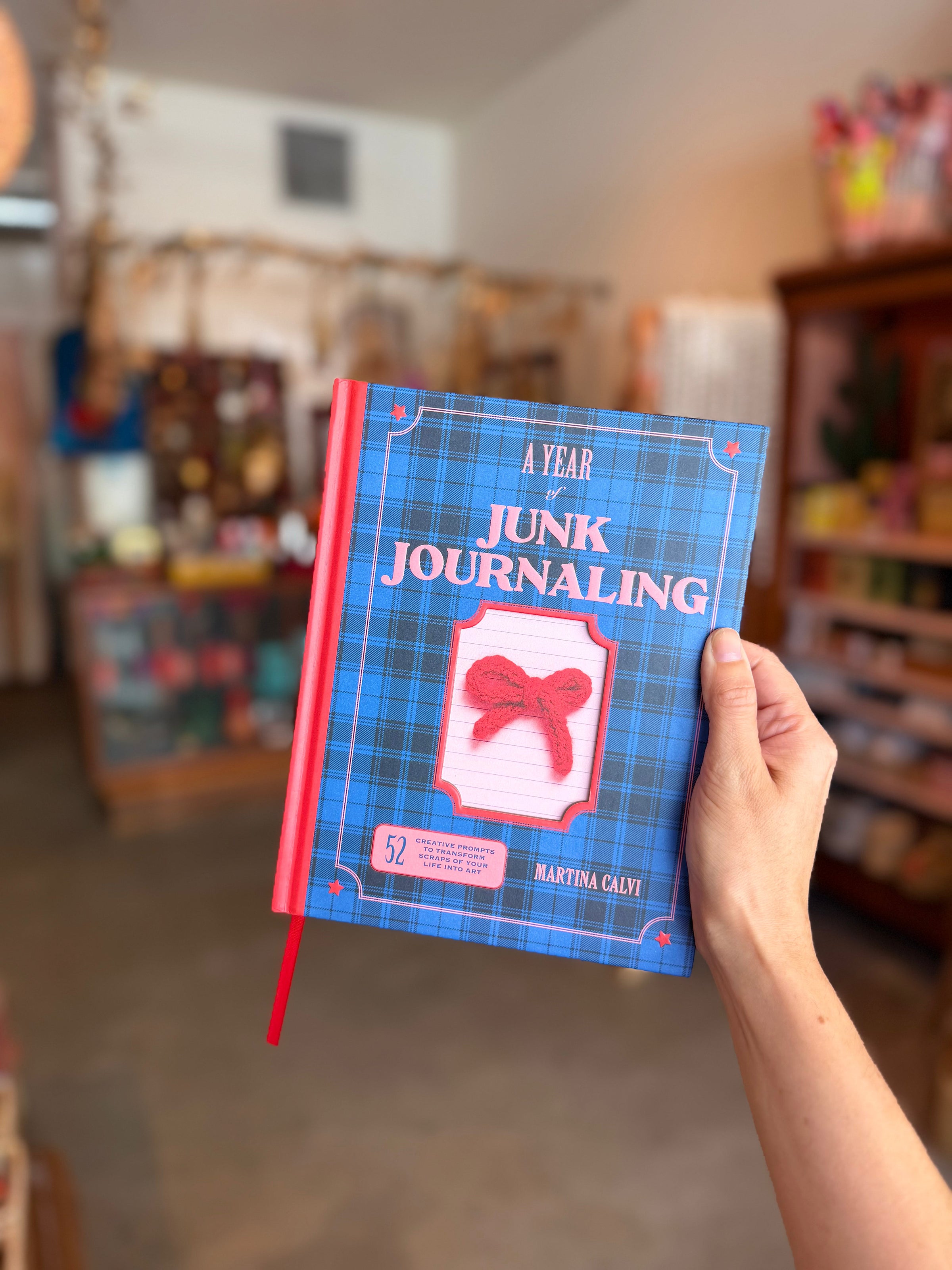 A hand holds a hardcover journal titled 'A Year of Junk Journaling' by Martina Cali. The cover features a blue plaid pattern with a pink and white bow graphic in the center, and a red spine and red ribbon bookmark.