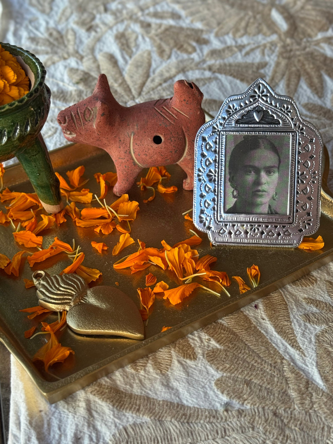 A styled still life featuring a silver-toned ornate portrait frame with a black-and-white photo of Frida Kahlo, alongside a terracotta figurine, a gold-toned heart-shaped object, and scattered orange marigold petals on a brass tray.