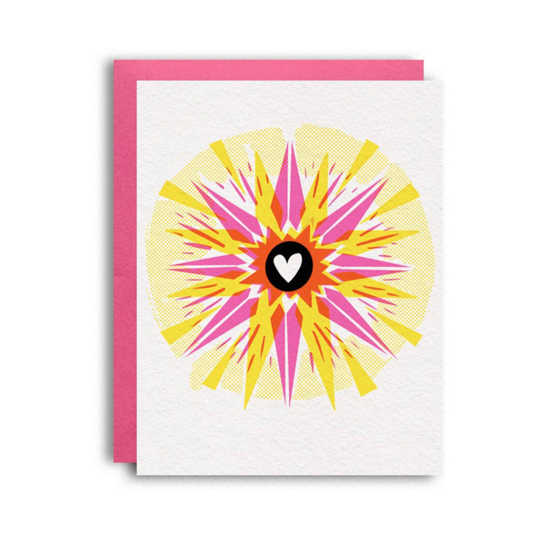 white card with a pink envelope featuring a yellow sunburst with a pink star and a black circle with a white heart in the center
