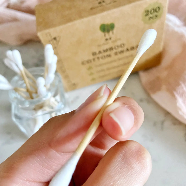 close up view of a hand holding a cootn swab with a box and jar of cotton swabs in the background.