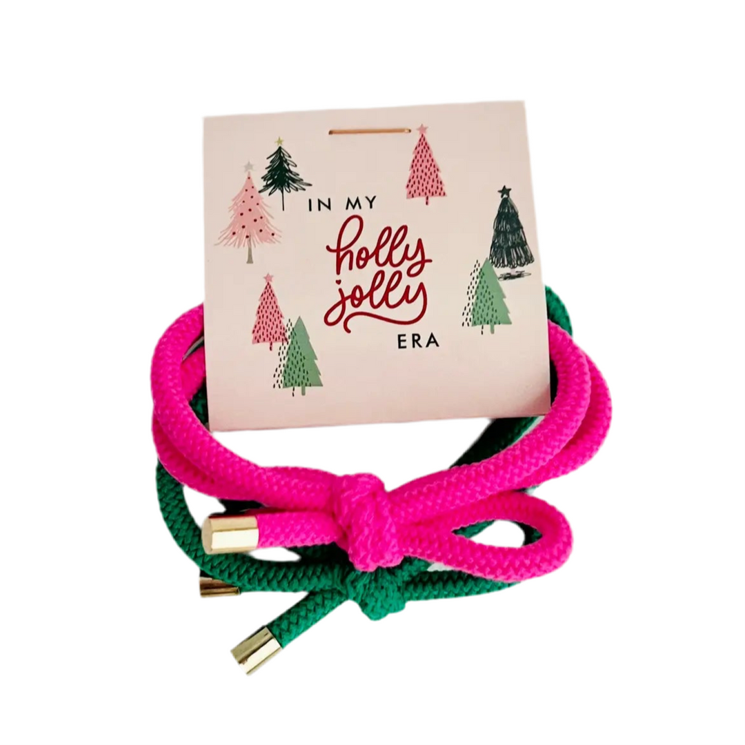 This image showcases a cute holiday-themed card with two vibrant knotted hair ties—one pink and one green—complementing the festive “In my holly jolly era” message on the card. 