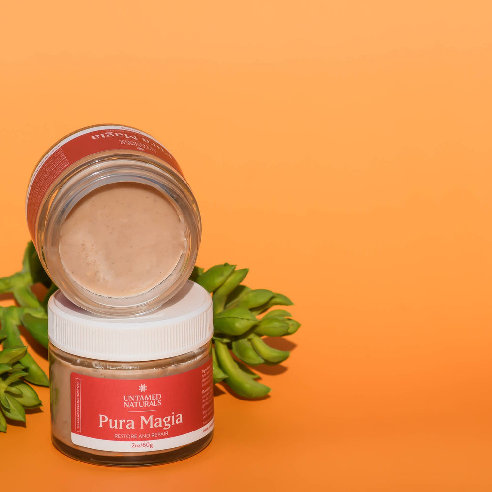 close up view of an open jar of pura magia moisturizer sitting on top of a closed jar of pura magia. Orange background with a succulent in the background. Brand: UnTamed Naturals