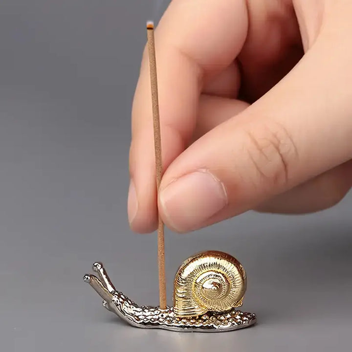 a gold and silver snail shaped incense holder with a hand placing an incense stick in it