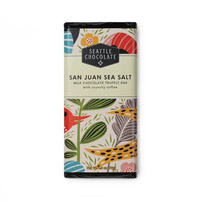 A Seattle Chocolate San Juan Sea Salt Milk Chocolate Truffle Bar in a colorful, nature-inspired wrapper featuring illustrated leaves, flowers, and a red bird. The packaging highlights the flavor combination of milk chocolate, crunchy toffee, and Pacific sea salt, with a net weight of 2.5 oz (70 g).