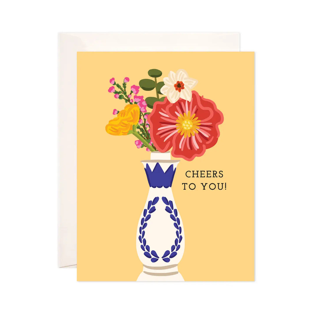 A greeting card with an illustration of a decorative blue and white vase holding a bouquet of colorful flowers against a warm yellow background. The text on the card reads "Cheers to You!" The card is paired with a matching white envelope.