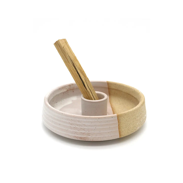 round ceramic tan and light pink palo santo burner features a hole in the center that is holding a palo santo stick.