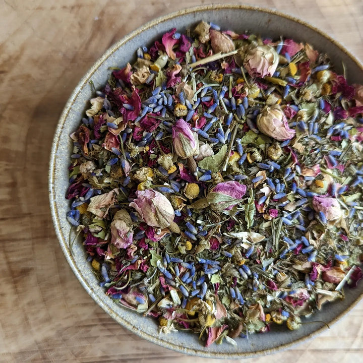 A close-up of a ceramic bowl filled with a colorful blend of dried herbs and flowers, including lavender, chamomile, rose petals, and other botanicals, used for making Serenity Herbal Tea.