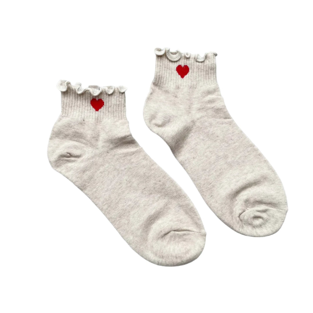 A pair of ivory-colored ankle socks with a small red embroidered heart near the ruffled trim, laid flat on a textured gray background.