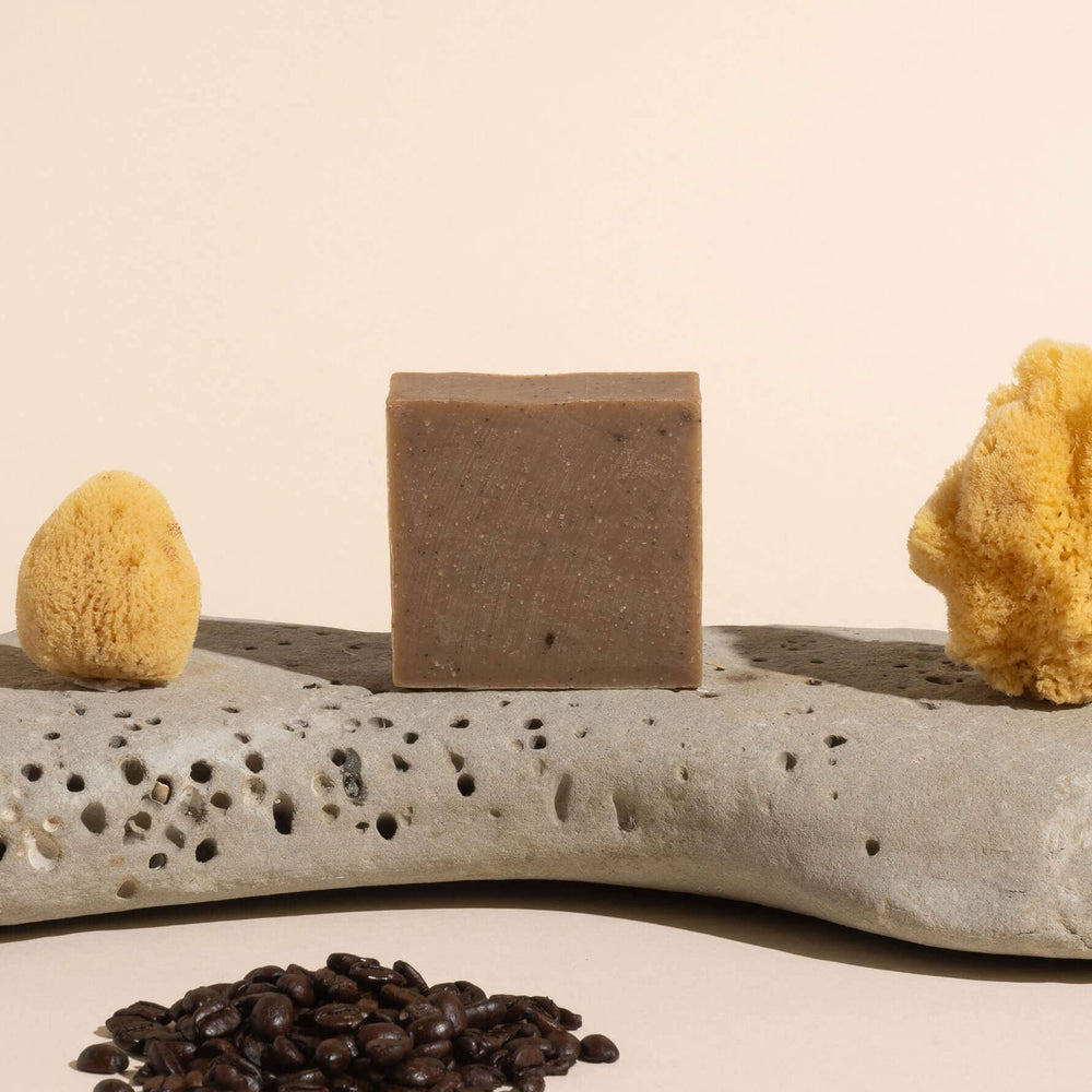 brown bar of soap on a gray stone surrounded by natural sponges and a pile of coffee grounds.