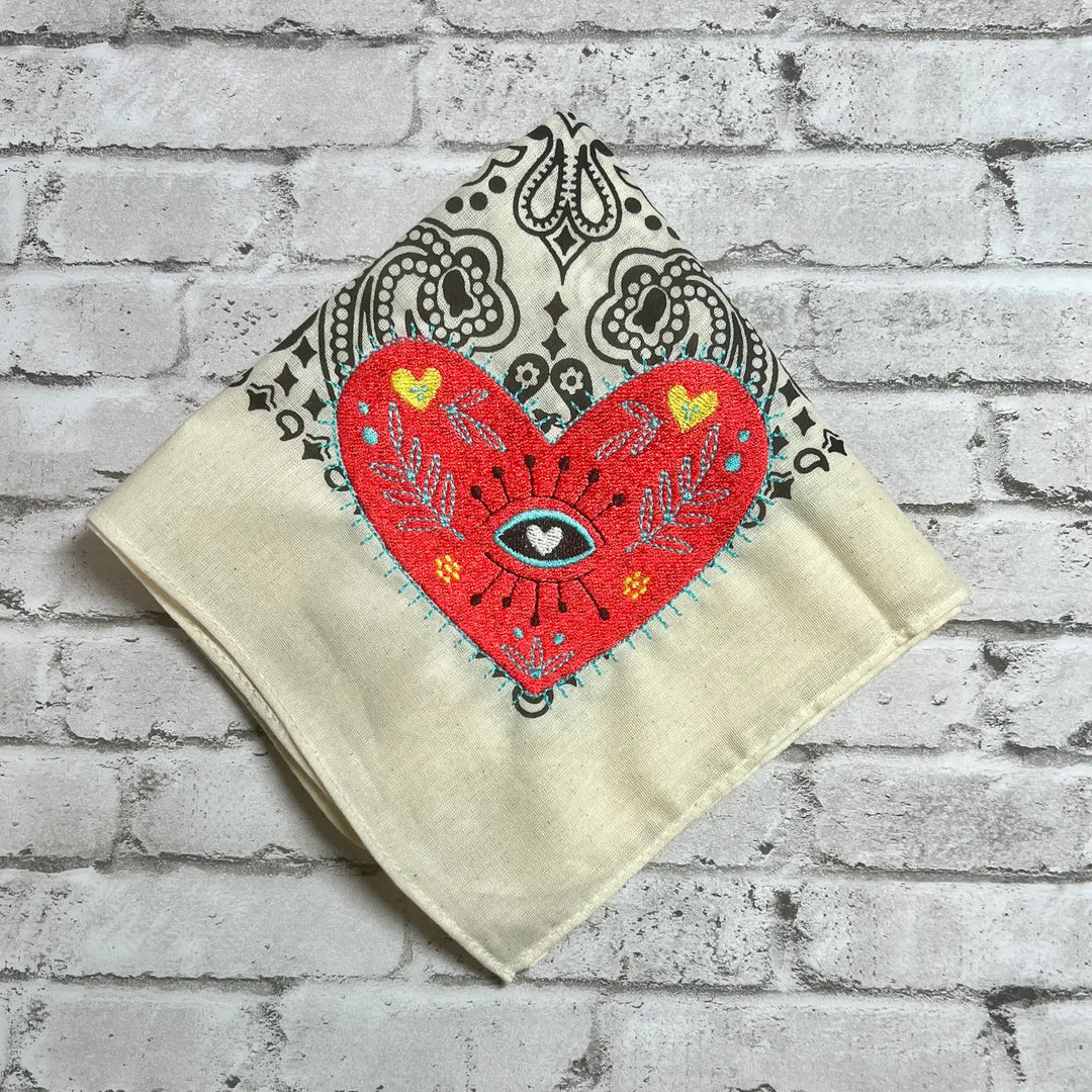 A Heart Embroidered Bandana with a red sacred heart, an all-seeing eye, and intricate embroidery on a beige fabric with a paisley pattern.