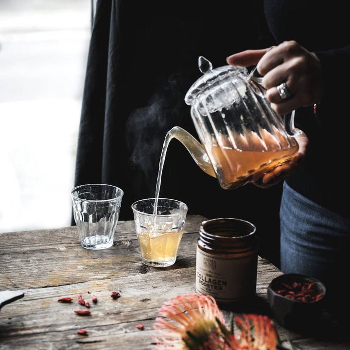 photo of a person pouring tea from a glass kettle into a glass cup with an open jar of collagen booster by the cup.