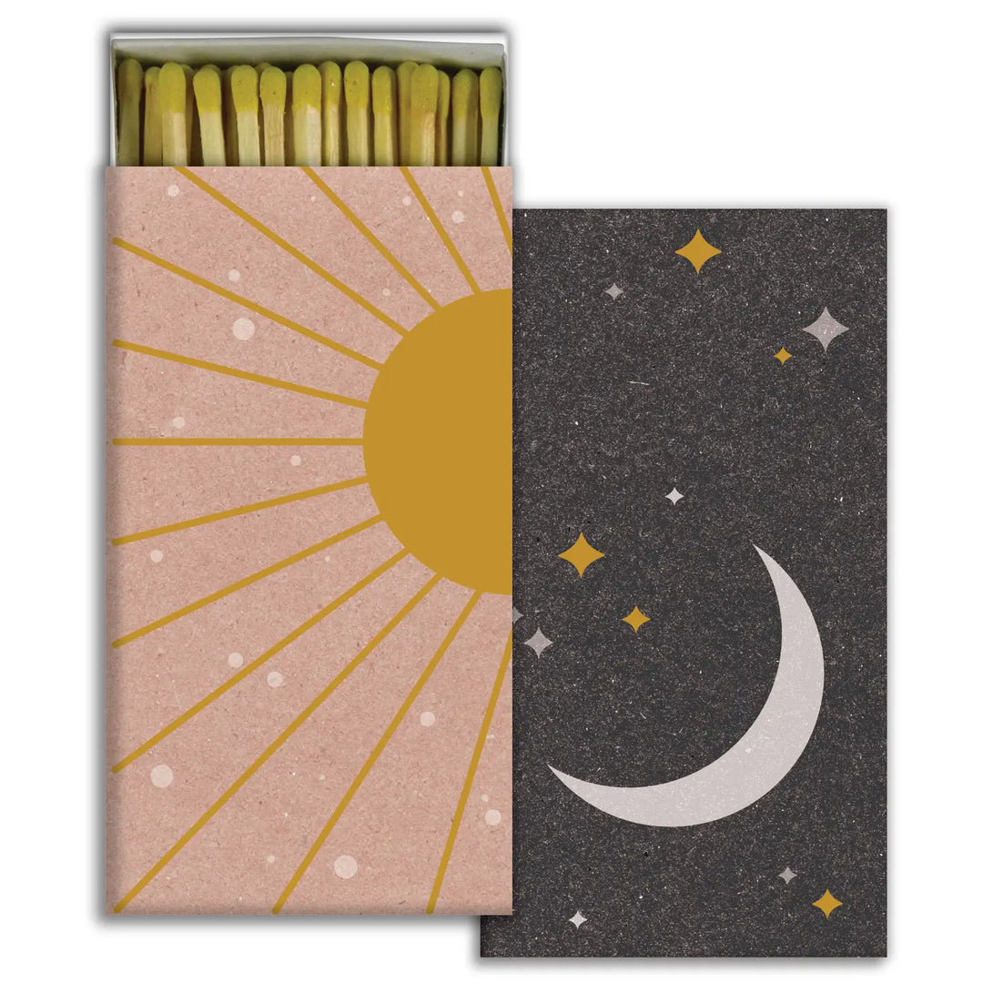 A stylish matchbox featuring two designs: one side has a warm, golden sun with rays against a textured beige background, and the other side showcases a crescent moon surrounded by stars on a dark, starry background. The matchbox is open, revealing long matches with yellow-tipped heads