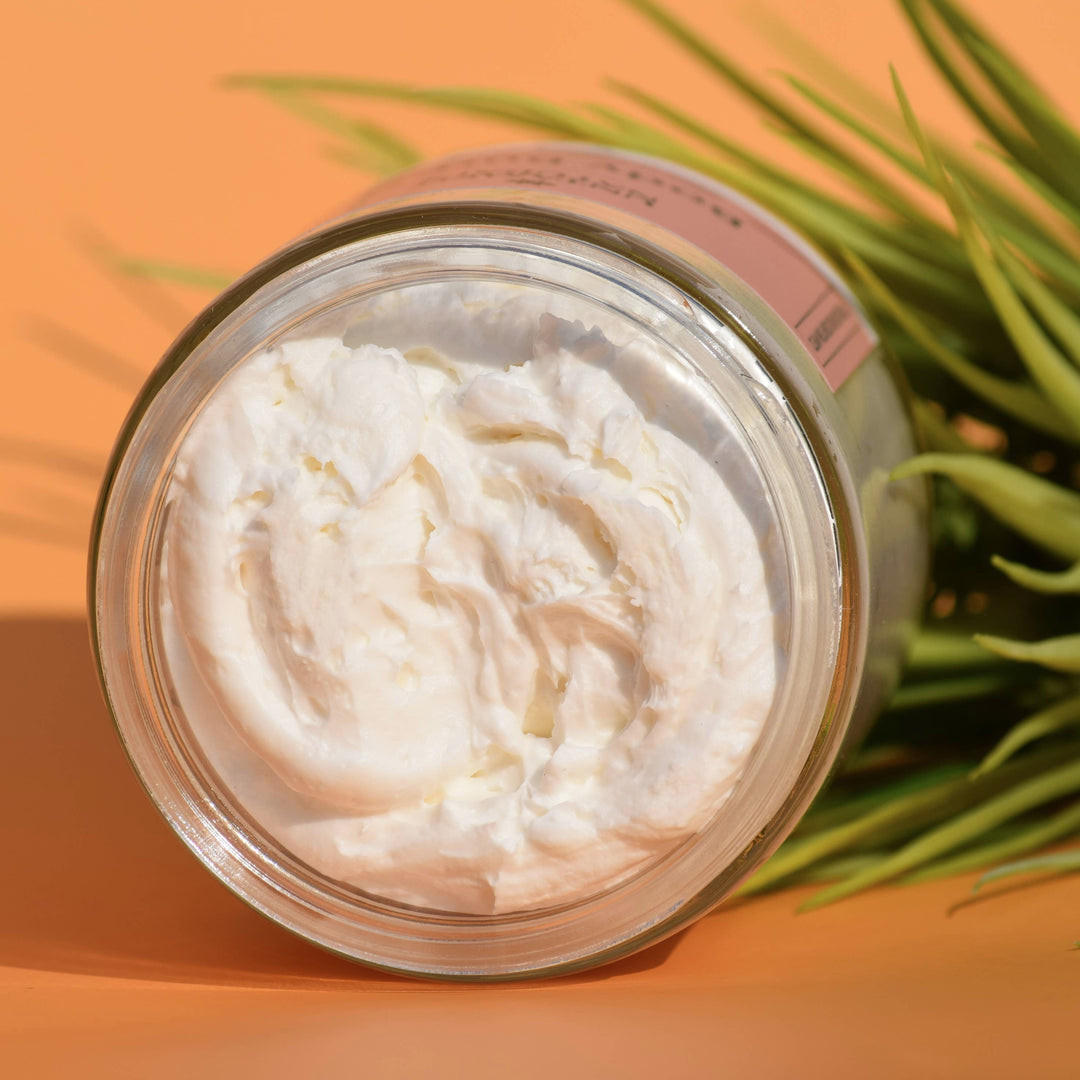 a clear open  jar of body butter on its side with an orange background and a plant. Brand: UnTamed Naturals