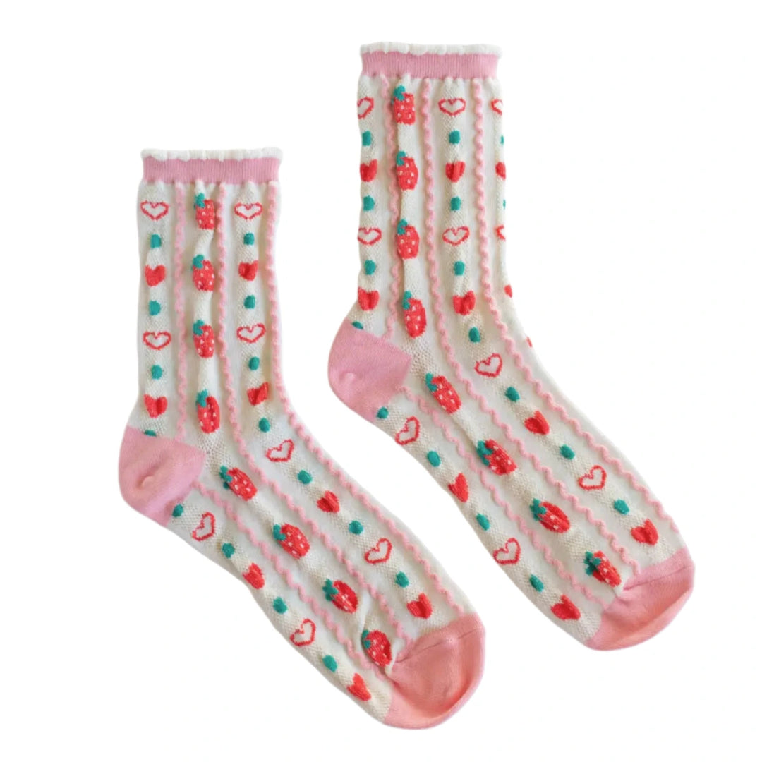 A pair of retro-inspired strawberry socks with an off-white background, decorated with red strawberries, blue-green dots, and red hearts. The socks have pink accents on the toe, heel, and ruffled trim, laid flat on a marble surface.