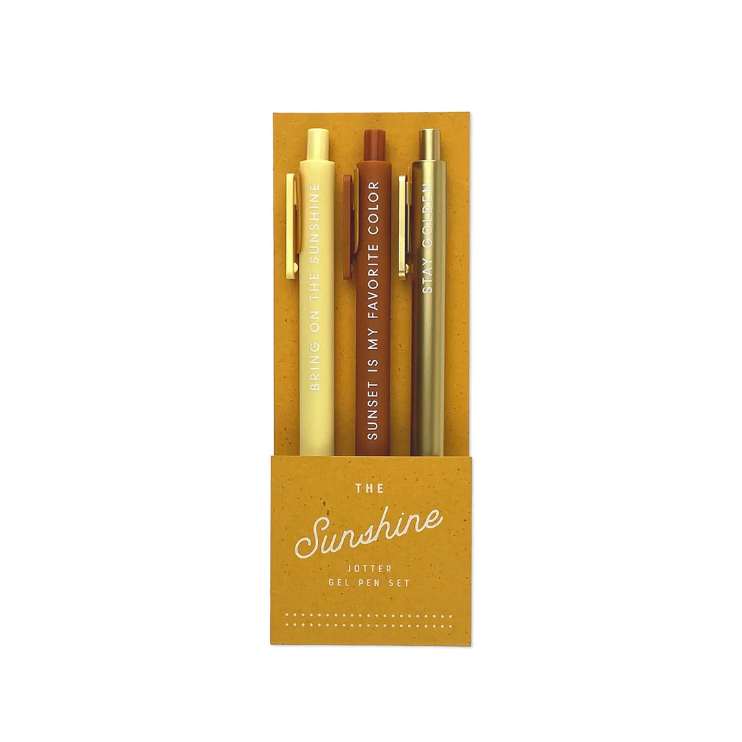 A set of three gel pens in a golden yellow packaging labeled "The Sunshine Jotter Gel Pen Set." The pens come in pastel yellow, rich burnt orange, and metallic gold, each featuring a cheerful phrase in white lettering: "Bring on the Sunshine," "Sunset is My Favorite Color," and "Stay Golden."