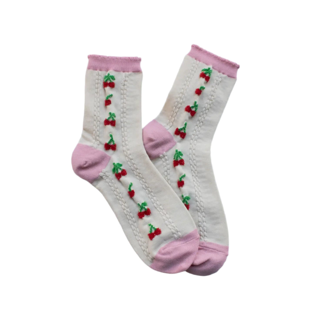 A pair of cream-colored socks with embroidered red cherries and green leaves running in vertical rows. The socks feature a cable-knit texture and soft pink accents on the toe, heel, and cuff, laid flat on a marble surface.