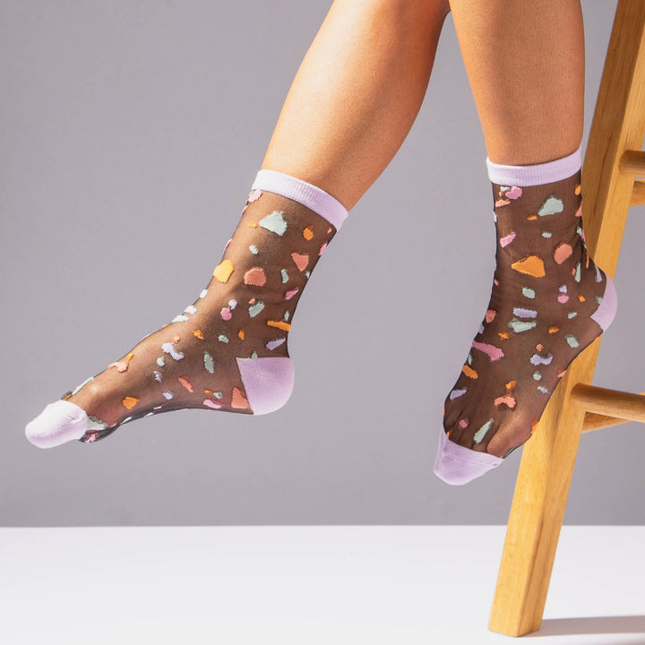 close up view of some feet wearing a pair of sheer socks with a dark gray background with multi-color fragments and matching lavender accents on toes and heels 