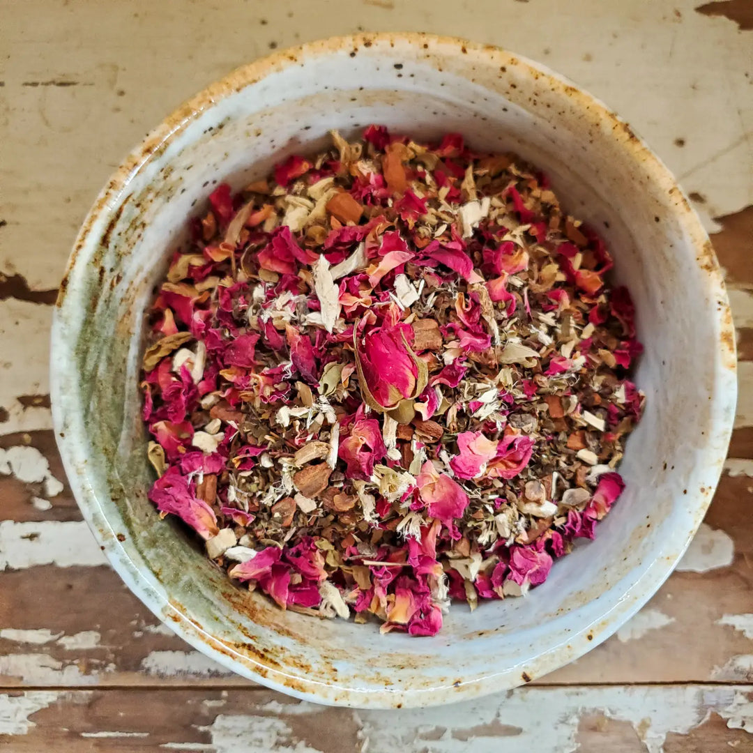 A rustic ceramic bowl filled with a loose herbal tea blend, featuring dried rose petals, cinnamon, marshmallow root, tulsi, and other botanicals. The bowl rests on a weathered wooden surface.