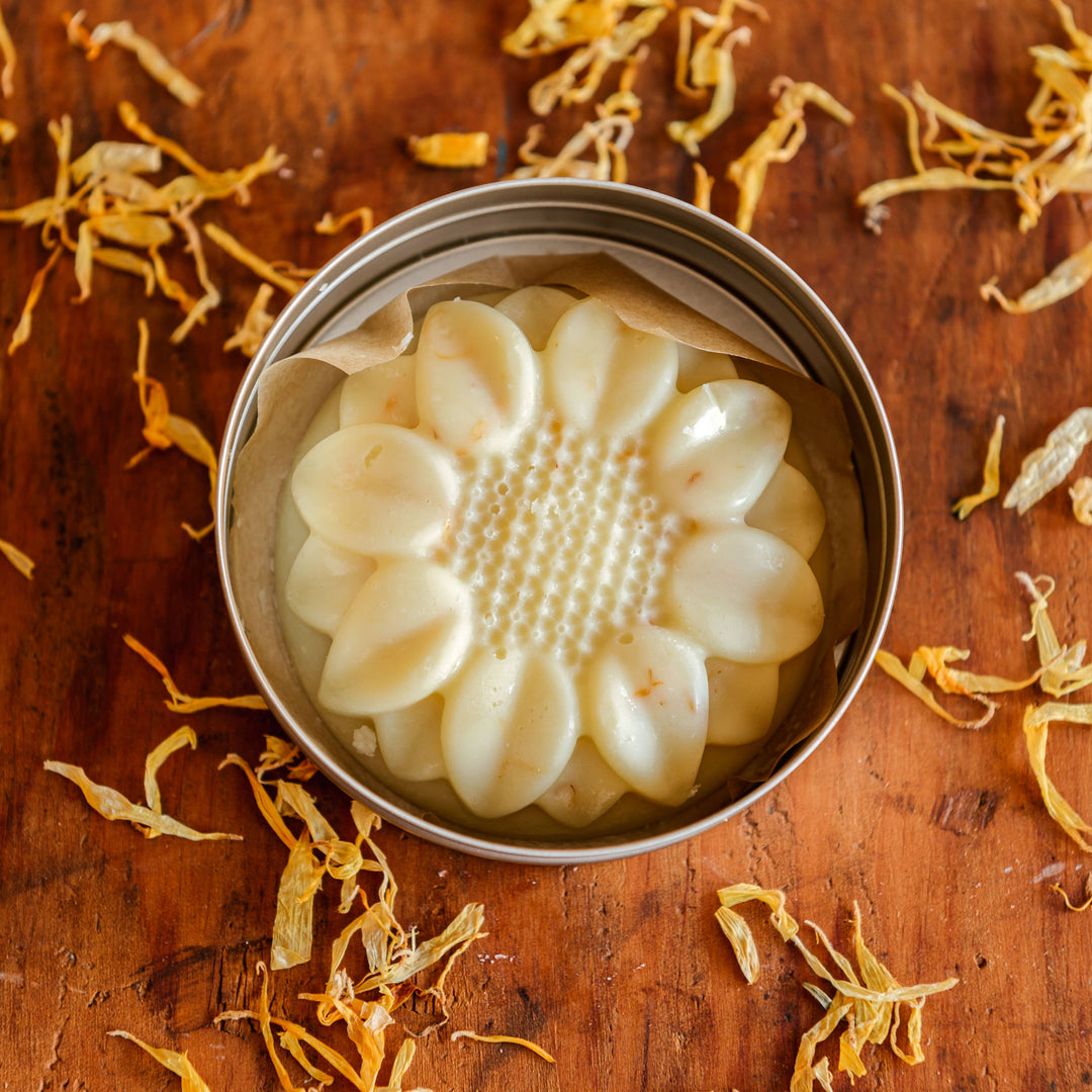 open tin with a flower shaped lotion bar on a wooden table with a sprinkle of dried calendula petals. Brand: Lovewild Design